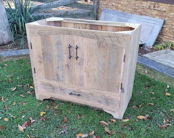Puede incluir: Un mueble de madera rústico con acabado natural. Cuenta con dos puertas con tiradores negros y un cajón debajo. El mueble está sobre una superficie de césped con hojas caídas.