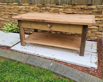 May include: A rustic wooden coffee table with a drawer and a lower shelf. The table is made of reclaimed wood and has a natural finish. It is sitting on a white surface with a green lawn in the background.