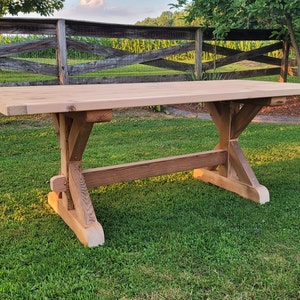 May include: A rustic wooden table with a natural finish and a trestle base. The table is set on a grassy lawn with a wooden fence in the background.