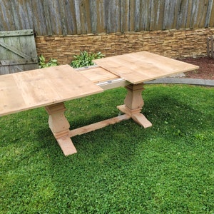 May include: A wooden table with a leaf extension. The table is made of light wood and has a simple design. The table is set up on a green lawn.