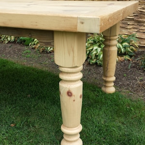 Puede incluir: Primer plano de una pata de mesa de madera con un dise&ntilde;o torneado. La pata de la mesa es de madera clara y tiene un acabado natural. La pata de la mesa est&aacute; sobre un c&eacute;sped verde.
