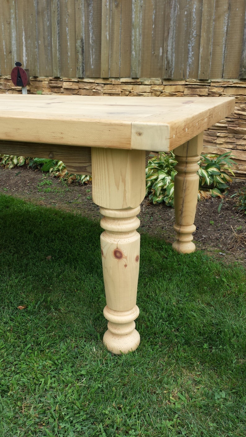 Puede incluir: Primer plano de una pata de mesa de madera con un dise&ntilde;o torneado. La pata de la mesa es de madera clara y tiene un acabado natural. La pata de la mesa est&aacute; sobre un c&eacute;sped verde.