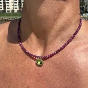 Puede incluir: Un collar que presenta una hilera de pequeñas cuentas facetadas de color magenta intenso. El collar incluye un único colgante circular con un marco dorado mate que rodea una piedra circular facetada de color verde lima brillante.