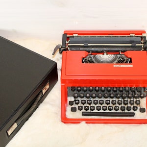 Peut inclure: Une machine à écrire Olivetti Lettera 32 rouge avec un étui noir. La machine à écrire a un clavier noir avec des touches blanches et un levier de retour chariot noir.