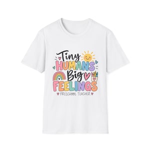 Può includere: Una t-shirt bianca a maniche corte con una stampa grafica colorata che recita Tiny HUMANS Big FEELINGS e PRESCHOOL TEACHER. Il design include un sole giallo, un arcobaleno, cuori e un contenitore di pastelli in tonalità pastello rosa, blu e arancione.
