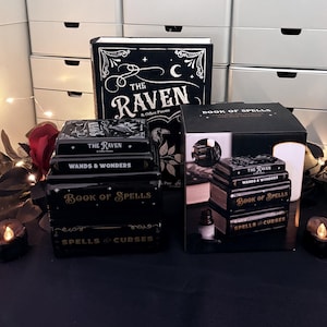May include: A black ceramic oil burner shaped like a stack of four books titled The Raven, Wands & Wonders, Book of Spells, and Spells & Curses. It is displayed next to its original black packaging box which features an image of the product.