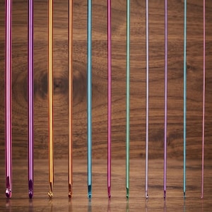 May include: A set of eleven colorful metal crochet hooks arranged in descending order of thickness. Each hook features a white label at the top indicating its size, ranging from 8.0 mm to 2.0 mm. The hooks are colored in shades of pink, purple, gold, orange, blue, and green.