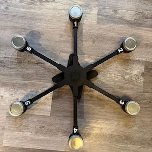May include: A black, star-shaped mounting bracket with six radiating arms, each numbered sequentially from one to six in white. At the end of each arm is a circular, silver-colored metal cap, creating a symmetrical, mechanical design.