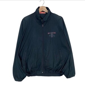 May include: A black long-sleeve zip-up jacket with a high collar and elastic cuffs. The garment features a small red and white brand tag at the inner neckline and a dark rectangular logo graphic on the upper left chest area with the text TRD.