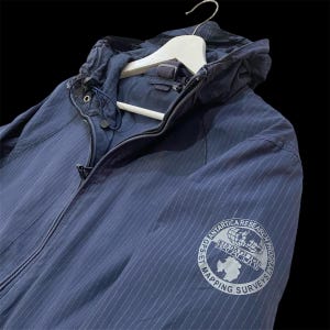 May include: A navy blue pinstriped hooded jacket with a full-length front zip and a drawstring hood. The left sleeve features a circular patch with the text NAPAPIJRI ANTARTICA RESEARCH PROGRAM GPS ET MAPPING SURVEYS and a globe graphic.