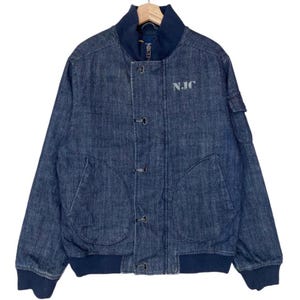 May include: A dark blue denim bomber jacket featuring a ribbed collar, cuffs, and hem. The front includes a vertical row of four metal toggle closures, two large side pockets, and the text N.JC embroidered in light gray on the upper chest area.