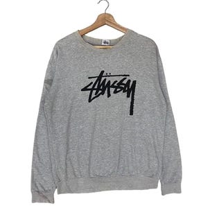 May include: A heather grey long-sleeve crewneck sweatshirt featuring a black Stüssy logo printed on the centre of the chest. The garment has a ribbed neckline, cuffs, and hem, and includes a small branded tag located inside the collar area.