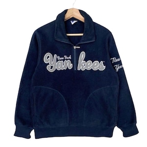 May include: A navy blue fleece pullover with a quarter-zip closure and a high collar. The front features the text New York Yankees in white and grey script lettering. The left sleeve also displays the text New York Yankees in white script.