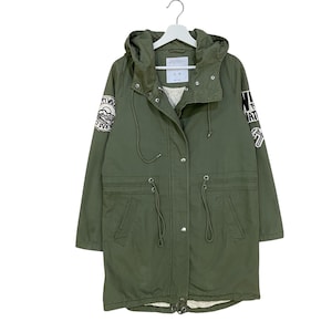 May include: An olive green hooded parka jacket featuring a front zip and snap button closure, a drawstring waist, and two front pockets. The sleeves are adorned with circular and rectangular patches with black and white graphic designs.