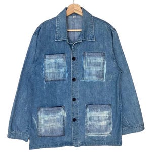 May include: A long-sleeve blue denim jacket featuring a classic collar, a button-down front with four dark buttons, and four large square patch pockets on the front. The pockets display a distinct lighter blue, horizontal brushstroke pattern.