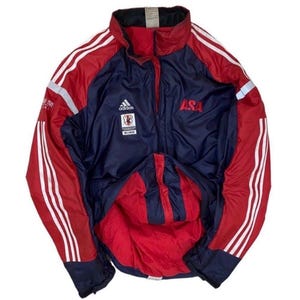 May include: A navy blue and red windbreaker jacket featuring white three-stripe detailing on the sleeves, a high collar, and a front zipper. The chest displays an Adidas logo, a small rectangular emblem, and the red text ASA on the right side.