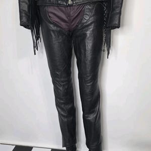 May include: A black leather motorcycle jacket with fringe details paired with black leather chaps worn over dark trousers and black lace-up boots. The jacket features a silver-tone front zipper, side buckles, and a chain accessory on the side.