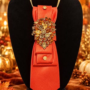May include: A bright orange necktie featuring a textured fabric, two small gold-toned buttons, and a large, ornate brooch centered on the front. The brooch is encrusted with numerous amber, yellow, and clear crystals in a floral arrangement.