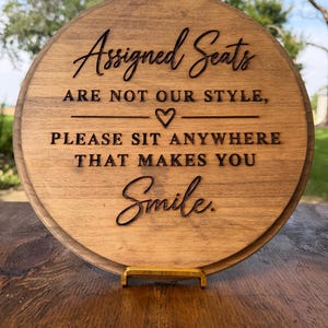 May include: A round, medium-brown wooden sign featuring dark, engraved text that reads: Assigned Seats ARE NOT OUR STYLE, a heart symbol, PLEASE SIT ANYWHERE THAT MAKES YOU Smile. The sign is supported by a small, gold-colored metal stand.