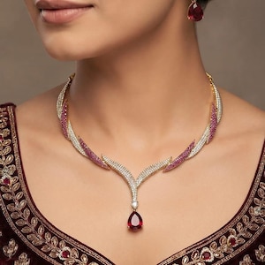 May include: A gold-toned necklace and matching earring set featuring pavé-set clear stones and deep red, pear-shaped center stones. The necklace has a curved, wing-like design with embedded red accents, while the earrings mirror the drop style.