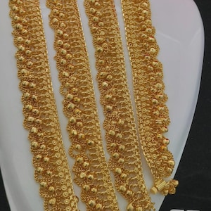 May include: A pair of gold-toned anklets featuring a multi-layered chain design with small spherical beads and intricate circular links. The anklets have a polished metallic finish and include a small clasp mechanism at the end of each chain.