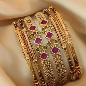 Peut inclure: Un ensemble de cinq bracelets rigides dorés avec des motifs texturés complexes. Deux bracelets présentent un design géométrique répétitif, tandis que les trois centraux sont reliés par un pont décoratif orné de pierres magenta carrées.
