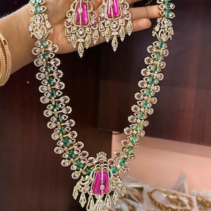 May include: A long necklace and matching drop earrings set featuring large, rectangular, vibrant pink stones as centerpieces, accented with smaller green stones and clear crystals set in a metallic frame with intricate, filigree-style detailing.