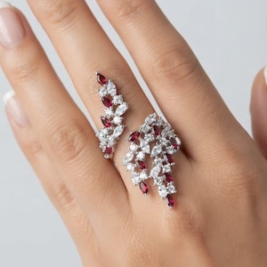 May include: An open-style ring featuring a silver-toned band adorned with marquise-cut clear white stones and deep red stones arranged in a cluster pattern that extends across the finger, creating a leaf-like or floral aesthetic design.