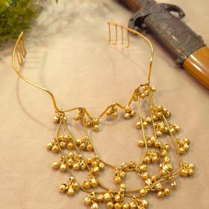 May include: A gold-toned metal headpiece featuring a thin, curved band with integrated combs at the ends. The front section is adorned with multiple draped gold chains, each embellished with numerous small, round, textured jingle bells.