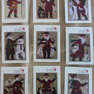 May include: Nine cross-stitch patterns featuring Santa Claus in various winter scenes. Each pattern includes the text "The Prairie Schooler" and a year, such as "2011 Santa" or "2022 Santa". The designs showcase Santa with animals and winter themes.