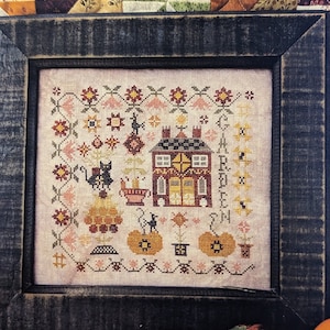 May include: A framed cross-stitch design featuring a house with a pumpkin patch and the text "Garden Home". The design is in a rustic style with a brown frame.
