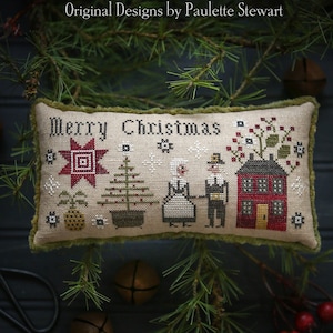 May include: A beige pillow with a green border features a cross-stitch design of a red star, a green Christmas tree, two figures in white and black clothing, and a red house with a white door. The pillow reads "Merry Christmas" in black lettering.