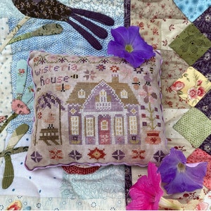 May include: A purple and white embroidered pillow with the text "Wisteria House" and a small house design. The pillow is on a patchwork quilt with floral patterns.