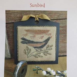 Puede incluir: Un patrón de punto de cruz de un pájaro sol en una rama con una puesta de sol en el fondo. El patrón está enmarcado en un marco negro y está sobre una superficie de madera. El texto "Threadwork Primitives" y "Sunbird" es visible en la imagen. El texto "Original Designs by Nan Lewis ~ Threadwork Primitives © 2016" y "Email: threadworkprimitives@verizon.net" también es visible en la imagen.