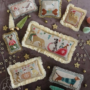May include: Six embroidered pillow ornaments featuring dogs in various holiday scenes. The pillows are decorated with gold and silver trim and are arranged on a brown background with gold stars and small artificial Christmas trees.