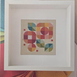 May include: A framed cross-stitch pattern titled "MOD BLOOM BOX" featuring a geometric floral design in shades of red, orange, yellow, and teal. The pattern is set against a neutral background and framed in a white square frame. The bottom of the image includes the text "ROBIN PICKENS CROSS STITCH PATTERNS."