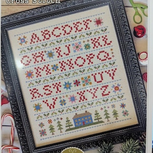 May include: A framed cross-stitch sampler titled "Home Town Holiday Sampler" with an alphabet design. The sampler features red, blue, green, and yellow cross-stitch letters and holiday-themed motifs. The frame is black with a decorative border. Designed by Lori Holt.