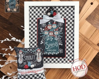 May include: A cross stitch pattern featuring a mason jar filled with snowmen and the text "Snowy Mason". The pattern is framed in a white frame with a black and white checkered background. The pattern is also available as a pillow with a black and white checkered fabric.