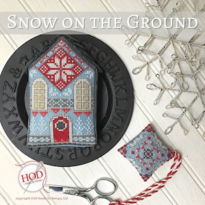 Peut inclure: Une décoration en point de croix représentant une maison bleue et rouge avec un motif de flocon de neige blanc. L'ornement est sur une assiette noire avec des lettres blanches qui épellent l'alphabet. Le texte "SNOW ON THE GROUND" est en haut de l'image.