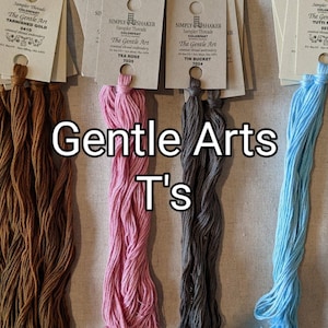 May include: Bundles of embroidery floss in shades of brown, pink, gray, and blue, with attached paper labels. The text "Gentle Arts T's" is overlaid on the image. The labels indicate the floss colors and brands.