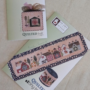 May include: Three cross-stitch pattern booklets with the Heart in Hand Needleart logo. One booklet is titled "Quilted Frill" and features a cross-stitched house design. Another is titled "Quilted Tiny Town" with a cross-stitched town scene and a matching pin cushion.