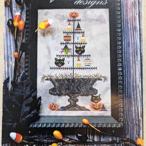 May include: A cross-stitch pattern for a Halloween feather tree. The design features a black cat, pumpkins, ghosts, and candy corn. The pattern is framed and displayed on a wooden background.