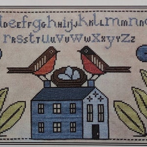 Può includere: Schema di punto croce con una casa blu con due uccelli rossi appollaiati sul tetto. Gli uccelli tengono un nido con tre uova bianche. L'alfabeto è ricamato sopra la casa e ci sono due piante verdi con fiori blu e bianchi su entrambi i lati.