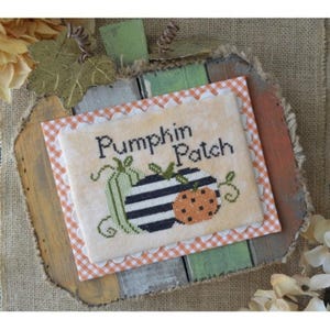 May include: A cross-stitch design featuring three pumpkins with the text "Pumpkin Patch" on a beige background. The design is framed with a white scalloped border and a peach and white gingham border.