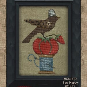 Puede incluir: Patrón de punto de cruz con un pájaro marrón posado sobre un tomate rojo encima de un carrete de hilo azul. El patrón se titula "Sew Happy" y forma parte de la colección "Another Itty Bitty Pattern".