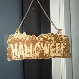 May include: A light-colored wooden hanging sign featuring the word HALLOWEEN in large, bold letters. The sign is decorated with laser-engraved illustrations of various horror movie characters and pumpkins, and it hangs from a piece of brown twine.