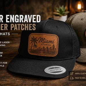 May include: A black mesh snapback hat featuring a rectangular tan leather patch with a laser-engraved Miami Florida skyline and palm tree design, positioned next to an olive green hat with a circular leather patch showing a mountain compass.