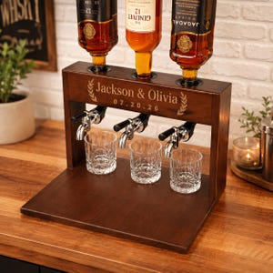 May include: A dark brown wooden spirit dispenser featuring three silver metal taps and three clear glass tumblers. The wood is engraved with the text Jackson & Olivia and the date 07.20.26, flanked by leaf motifs. Three glass bottles are inverted.