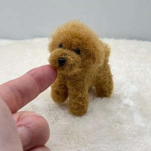 以下が含まれることがあります： ふわふわとした質感の金茶色の羊毛フェルトで作られた小さな犬のフィギュア。暗いビーズの目と小さな黒い鼻が特徴です。4本の足で立ち、丸い頭と垂れた耳を持つこのミニチュア彫刻は、羊毛繊維を使用して丁寧に作られています。