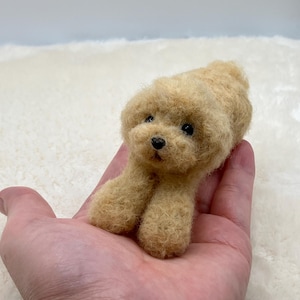 以下が含まれることがあります： カールした質感のウールの毛並みを持つ、明るい茶色の犬の小さなニードルフェルト彫刻。2つの暗い丸い目と小さな黒い三角形の鼻が特徴で、前足を前に伸ばして横たわっている姿勢で表現されています。